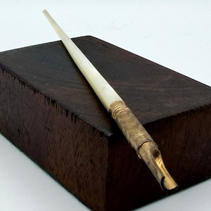 May include: A dip pen featuring a tapered, off-white handle and a metallic gold-toned nib holder with fine ribbed detailing. The pen is positioned diagonally across a dark brown, rectangular wooden block with a visible wood grain texture.