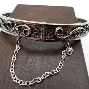 Puede incluir: Brazalete rígido plateado con bisagras y diseños intrincados de filigrana y calados. El brazalete tiene una cadena de seguridad y se muestra sobre una superficie de madera oscura. La filigrana incluye patrones florales y geométricos.