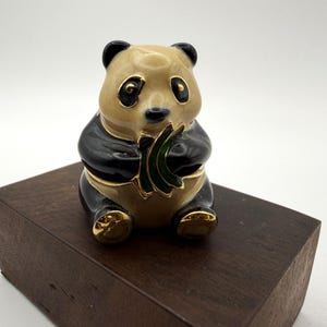 May include: A ceramic panda figurine with black and tan fur, black eyes, and gold detailing. The panda is holding green bamboo and is sat on a dark brown wooden block.