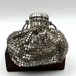 May include: A silver-toned metal mesh pouch featuring a flexible, interlocking square-link construction. The bag has a circular, engraved metal top frame with a series of small eyelets and two side rings for attachment, creating a gathered effect.