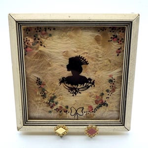 Könnte beinhalten: Gerahmte Silhouette einer Frau in Schwarz, verziert mit einem dekorativen Kopfschmuck und einem Blumengirlande. Der cremefarbene Rahmen hat eine schwarze Umrandung. Zwei kleine, goldfarbene Ohrringe mit rautenförmigen Zentren sind im Vordergrund.