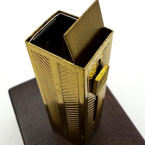 May include: A vintage gold-toned metal cigarette case with a rectangular shape and a hinged lid. The case features a geometric design and a small latch on the side. It sits on a dark wooden surface.