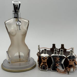 May include: A translucent, torso-shaped perfume bottle with a silver spray nozzle and a small circular tag reading TIRER PULL, positioned next to a bracelet featuring multiple miniature metallic torso charms connected by thin, light-colored cord.