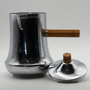Puede incluir: Una cafetera cromada con asa y tapa marrones. La cafetera tiene una base acampanada y un pico. La tapa es redonda con un pomo marrón. El asa es de material marrón con un diseño acanalado.