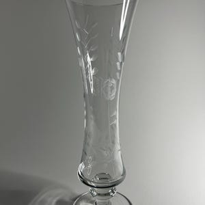 Könnte beinhalten: Ein klares, geätztes Champagnerglas mit einem breiten, ausgestellten oberen Teil und einem runden Fuß. Das Glas weist ein zartes Blumen- und Blättermuster auf. Der Stiel ist kurz und hat ein dekoratives Design.