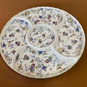 May include: A white ceramic serving platter with five sections, decorated with a floral pattern in blue, purple, yellow, and green. The platter has a scalloped edge with a green trim, suitable for serving appetizers or snacks.
