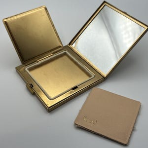 May include: Open gold-toned compact with a mirror and a powder compartment. A separate, square, tan powder puff with the word "Vincent" is next to the compact. The compact is open, revealing the mirror and powder well.