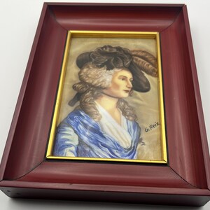 May include: A framed portrait of a woman in a hat with feathers, wearing a blue shawl. The artwork is in a gold frame, set within a dark red wooden frame. The artist's signature, "G. Reiz," is visible in the lower right corner.