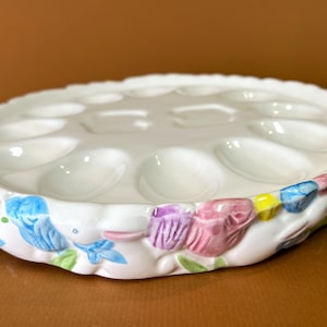 Pode incluir: Suporte para ovos de cerâmica branca com borda ondulada. A borda é decorada com desenhos florais coloridos em azul, rosa, amarelo e verde. A bandeja de ovos tem múltiplas reentrâncias circulares.