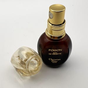 May include: A brown Poison Eau de Toilette perfume bottle with a gold-coloured cap and accents. The clear cap is off to the side. The Christian Dior logo is visible on the bottle.