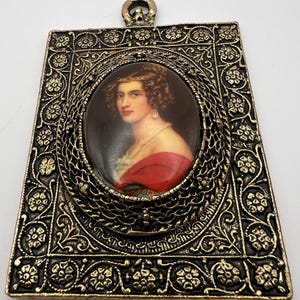 May include: An antique pendant featuring a portrait of a woman in a red shawl. The oval portrait is set within a filigree frame, surrounded by a rectangular, ornate, gold-toned metal border. A small loop is at the top for hanging.