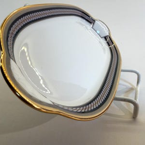 May include: A white, heart-shaped ceramic dish with a gold rim and a decorative border of black, gray, and gold. The dish is displayed on a white metal stand, showcasing its elegant design. This decorative dish is perfect for serving small portions.
