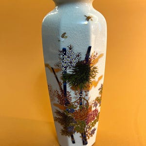 May include: A tall, off-white ceramic vase with a gold rim and a hexagonal base. The vase features a detailed floral and bamboo design in shades of green, orange, blue, and gold. The surface has a crackle glaze.