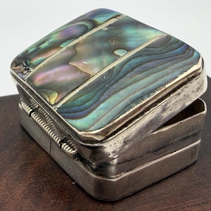 May include: A small rectangular box with a hinged lid featuring inlaid iridescent abalone shell panels in shades of blue, green, and purple. The box has a metallic silver-toned frame with a visible hinge mechanism on the back side.