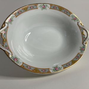 May include: Oval white serving bowl with gold trim and handles. The rim features a decorative border with pink roses and a blue and gold lattice pattern. A classic piece for serving.