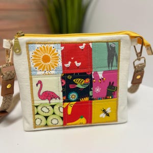 May include: A cream-colored crossbody bag with a patchwork design. The patchwork features a sunflower, flamingo, kiwi slices, birds, a sloth, a llama, a bee, and bananas. The bag has a yellow zipper and gold-colored hardware.
