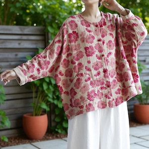 May include: A beige and pink floral print jacket with a round neckline, button closure, and two front pockets. The jacket has long sleeves with beige cuffs. The model is wearing white trousers.