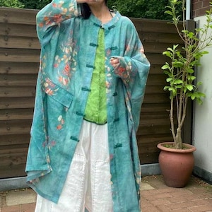 May include: A long, flowing teal jacket with a floral pattern in shades of orange and pink. The jacket has a mandarin collar and green frog closures. Underneath, a lime green top and white trousers are visible, creating a layered look.