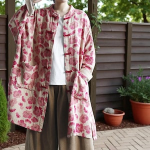 May include: A long, open-front jacket with a floral pattern in shades of pink on a cream background. The jacket features large pockets, red toggle closures, and a mandarin collar. The sleeves are long, and the jacket is worn over a white top and brown pants.