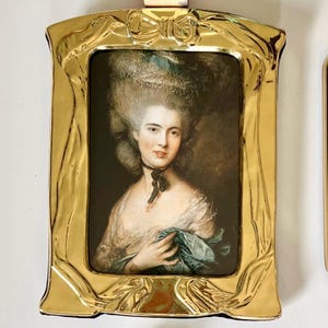 May include: A rectangular portrait frame with a polished gold finish, featuring a molded bow detail at the top and flowing ribbon-like edges. The frame contains a print of an 18th-century style painting of a person with an elaborate hairstyle.