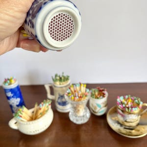 May include: A collection of small ceramic and glass containers filled with colorful wooden matches. One blue and white patterned container is held up to show a circular brown strike pad with a white honeycomb texture on its bottom surface.