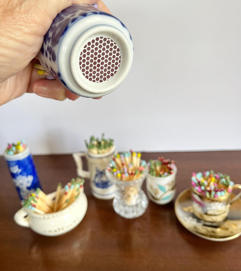 May include: A collection of small ceramic and glass containers filled with colorful wooden matches. One blue and white patterned container is held up to show a circular brown strike pad with a white honeycomb texture on its bottom surface.