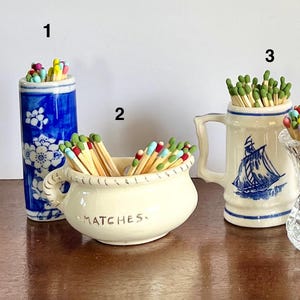 May include: Three ceramic containers holding wooden matches. Container 1 is a tall blue and white floral cylinder. Container 2 is a cream-colored bowl with the word MATCHES printed on the front. Container 3 is a cream-colored mug with a blue sailboat.
