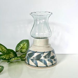 May include: A ceramic oil lamp base with a cream-colored glaze and a hand-painted blue leaf pattern around the bottom. The lamp features a clear, scalloped-edge glass chimney top that sits securely on the wide, rounded ceramic base.