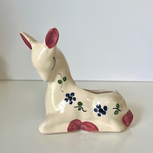 May include: A cream-colored ceramic deer figurine planter with a glossy finish. The deer is in a reclining position with its head bowed. It features painted pink ears, blue floral accents with green stems, and a pink tail detail on its back.