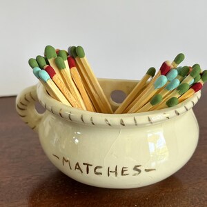 May include: A cream-colored ceramic pot with a handle and a rope-textured rim, featuring the word MATCHES printed in brown on the front. The pot is filled with wooden matches that have colorful tips in shades of green, red, and light blue.
