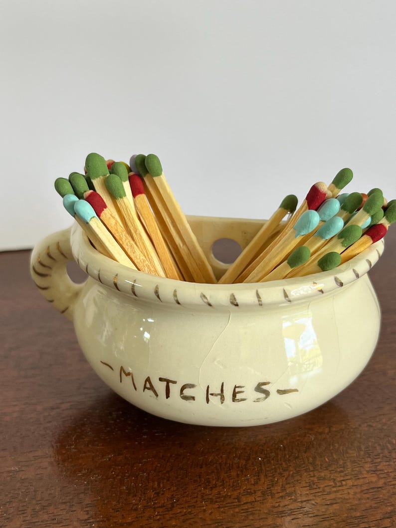 May include: A cream-colored ceramic pot with a handle and a rope-textured rim, featuring the word MATCHES printed in brown on the front. The pot is filled with wooden matches that have colorful tips in shades of green, red, and light blue.