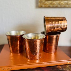 May include: A set of three hammered copper cups with a metallic finish. Two cups sit upright on a flat surface, while a third cup is positioned horizontally on top of a fourth cup, which is also standing upright. The cups have a cylindrical shape.