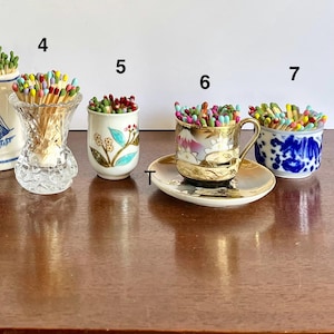 May include: Four decorative containers labeled 4, 5, 6, and 7, each filled with multicolored matches. Container 4 is a clear cut-glass vase, 5 is a white ceramic cup with floral motifs, 6 is a gold-rimmed teacup on a saucer, and 7 is a blue and white bowl.