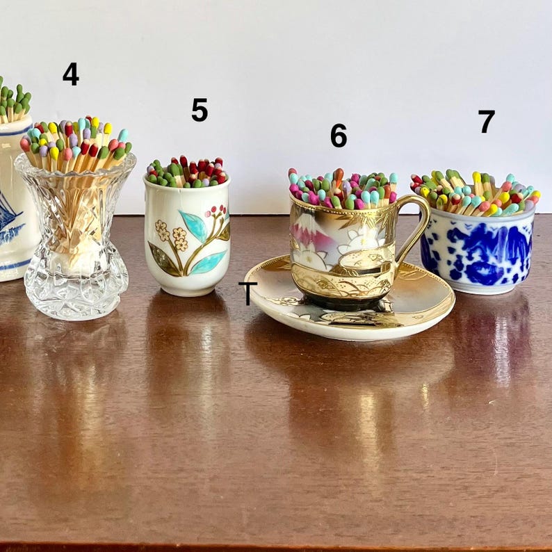 May include: Four decorative containers labeled 4, 5, 6, and 7, each filled with multicolored matches. Container 4 is a clear cut-glass vase, 5 is a white ceramic cup with floral motifs, 6 is a gold-rimmed teacup on a saucer, and 7 is a blue and white bowl.