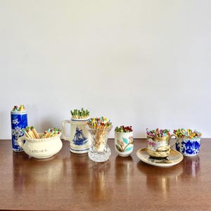 May include: A collection of six decorative containers filled with colorful matches. The set includes a blue and white floral cylinder, a cream bowl labeled MATCHES, a white mug with a blue ship, a clear glass vase, a floral cup, and a teacup.