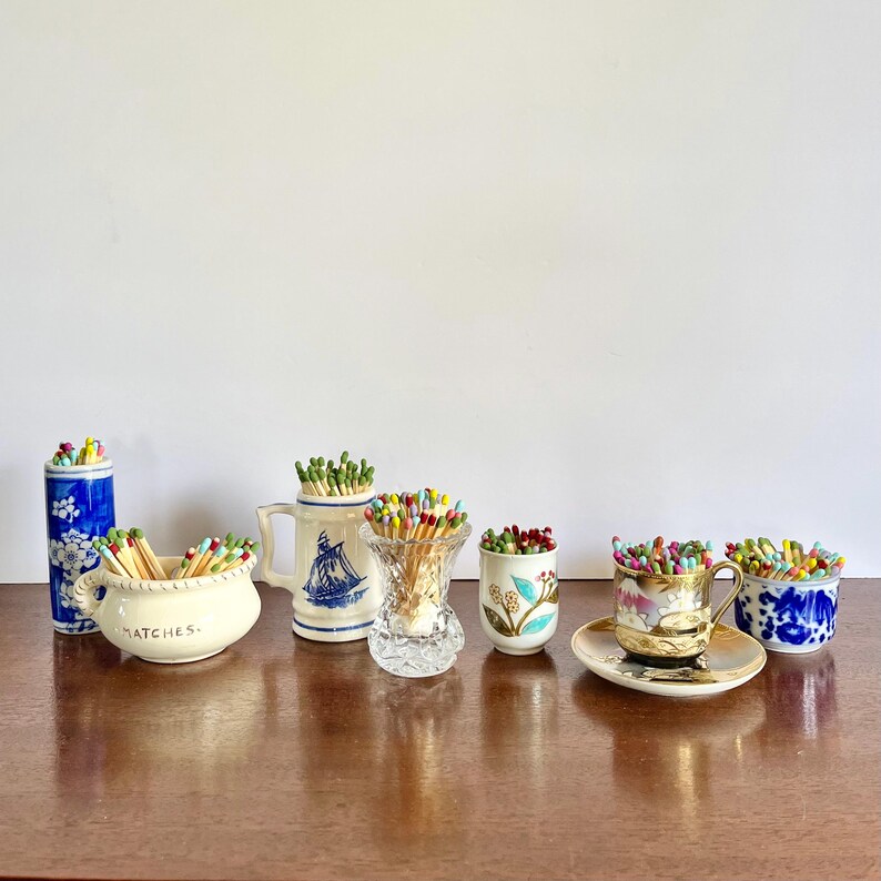 May include: A collection of six decorative containers filled with colorful matches. The set includes a blue and white floral cylinder, a cream bowl labeled MATCHES, a white mug with a blue ship, a clear glass vase, a floral cup, and a teacup.