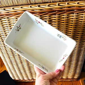 May include: A rectangular white ceramic baking dish with floral corner accents in pink, blue, and green. The dish is held at an angle, showcasing its clean lines and decorative details. The background is a woven wicker basket.