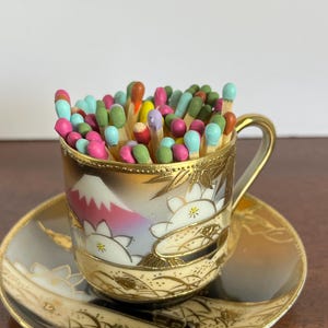 May include: A small porcelain teacup and saucer set featuring a painted mountain and floral design with gold trim, filled with a collection of wooden matches with colorful painted tips in shades of pink, green, blue, orange, and yellow.