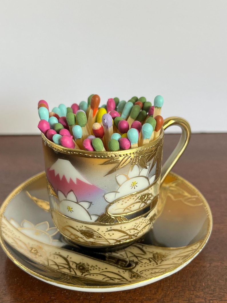 May include: A small porcelain teacup and saucer set featuring a painted mountain and floral design with gold trim, filled with a collection of wooden matches with colorful painted tips in shades of pink, green, blue, orange, and yellow.
