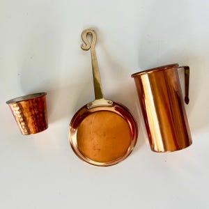 May include: Three copper kitchenware items: a small hammered cup, a small frying pan with a brass handle, and a tall pitcher with a handle. The items are arranged on a white surface. The copper has a shiny, reflective surface.