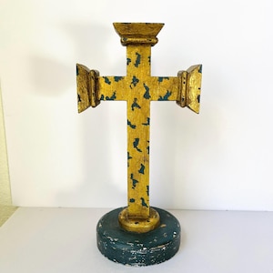 May include: A wooden cross with a gold finish and blue accents. The cross is mounted on a circular base painted in teal. The cross has a decorative design and is likely used for religious purposes.