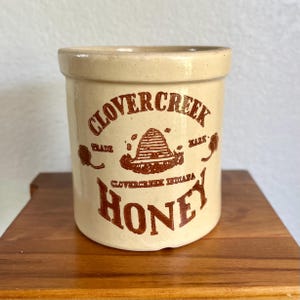 May include: A cylindrical beige ceramic crock featuring brown printed text and graphics. The front displays the words CLOVERCREEK HONEY, a beehive illustration, the text TRADE MARK, and the location CLOVERCREEK INDIANA with small clover icons.