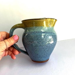 May include: A handmade ceramic pitcher with a blue, green, and yellow glaze. The pitcher has a rounded body, a curved handle, and a spout. The pitcher is held by a hand with pink nail polish.