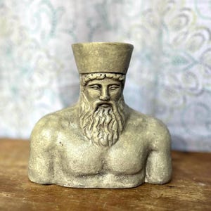 May include: A beige bust of a bearded man wearing a hat. The sculpture has a detailed face and chest, with a long, flowing beard. The bust is made of a stone-like material and sits on a wooden surface.