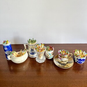 May include: A collection of ceramic and glass containers filled with colorful matches. The set includes a blue and white floral cylinder, a cream-colored bowl labeled MATCHES, a white mug with a blue ship, a clear glass vase, a floral cup, and a teacup.