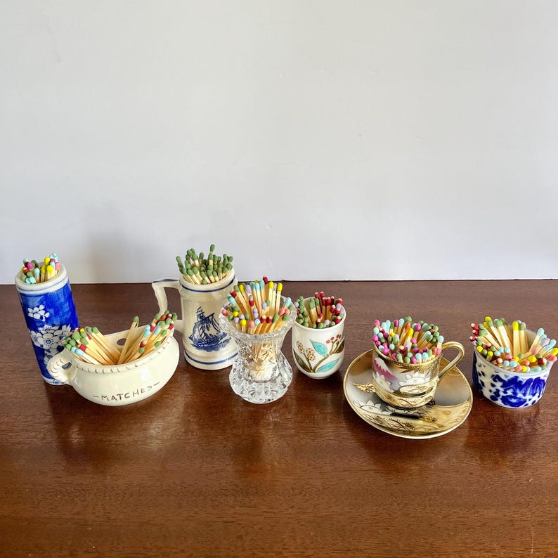 May include: A collection of ceramic and glass containers filled with colorful matches. The set includes a blue and white floral cylinder, a cream-colored bowl labeled MATCHES, a white mug with a blue ship, a clear glass vase, a floral cup, and a teacup.