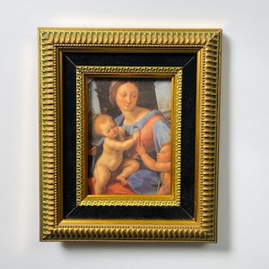 May include: A framed print of a Renaissance-style painting. The artwork depicts a woman in blue and red robes with two children. The frame is gold with a black inner border, and the overall aesthetic is classic and elegant.