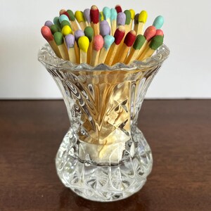 May include: A clear, faceted glass vessel containing a collection of wooden matches with multi-colored heads. The match heads are painted in various shades including yellow, pink, blue, green, red, and purple, contrasting with the clear glass.
