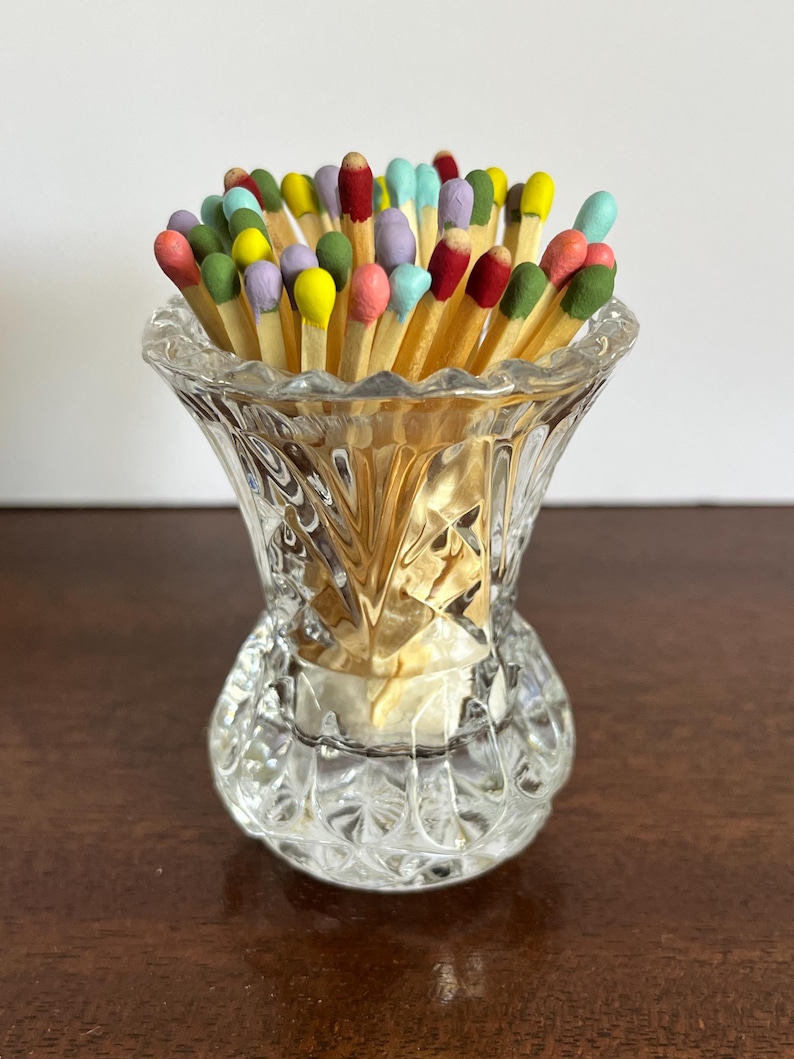 May include: A clear, faceted glass vessel containing a collection of wooden matches with multi-colored heads. The match heads are painted in various shades including yellow, pink, blue, green, red, and purple, contrasting with the clear glass.