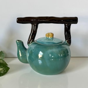 May include: A teal ceramic teapot with a brown handle and accents. The teapot has a round body, a spout, and a lid with a small gold-colored knob. The handle is designed to look like a branch.